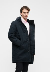 Fekete kapucnis parka, cipzáros elülső résszel, oldalsó zsebekkel és gombdíszítéssel. Simaszerkezetű, közepes hosszúságú, laza fazonú, rózsaszín póló felett.