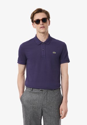 Paarse polo shirt van katoenen stof met een kraag en een knoopsluiting met twee knopen, voorzien van een klein groen geborduurd logo. Grijze gestreepte broek.