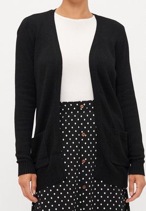 Vue recadrée d’un mannequin portant un cardigan noir ouvert sur le devant par-dessus un haut blanc et une jupe noire à pois blancs avec des boutons marron.