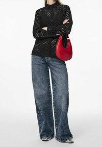 Černá košile s knoflíky a výřezovými detaily, spárovaná s širokými modrými džíny a zakřivenou červenou kabelkou. Metalické boty dokončují vzhled.