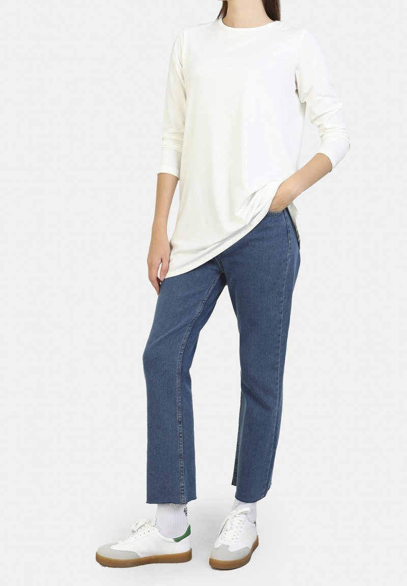 Wit top met lange mouwen gecombineerd met blauwe jeans met rechte pijpen en witte sneakers met een groene accent. Glad materiaal en een ontspannen pasvorm.