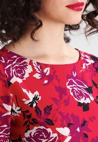 Nainen, jolla on kihartuneet tummat hiukset, pukeutuu punaisiin kukallisiin mekkoihin, joissa on purppuran-, valkoisen- ja mustanvärisiä kukkia, sekä kirkasta punaista huulipunaa.