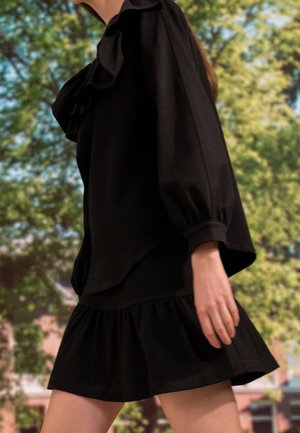 Woman walking outdoors wearing a black long-sleeve blouse with ruffles and a short black skirt with a ruffled hem.