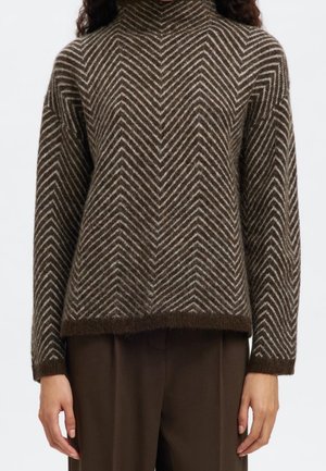 Femme portant un pull en maille à motif chevron marron et blanc avec des manches longues et un pantalon marron foncé.