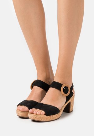 Fødder iført sorte ruskinds sandaler med åbne tæer, korkblok hæle og store runde flettede spænder mod en hvid baggrund.