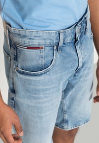 Světlé modré denimové šortky s předními a zadními kapsami, které nosí osoba v světlém modrém tričku, odhalující část jejich rukou a horních nohou.