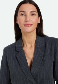 Blazer a righe scure con colletto a giogo e vestibilità sartoriale. Il tessuto ha una texture liscia, con sottili strisce bianche a contrasto.