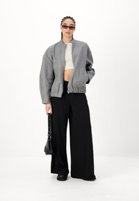 Sivi bomber s patentnim zatvaračem, spuštenim ramenima i rebrastim rubom, u kombinaciji s crnim hlačama širokog kroja i elegantnim sunčanim naočalama.