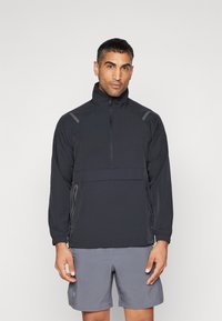 Zwarte pullover jack met een hoge kraag, halve rits, elastische manchetten en een borstzak. Gedeeltelijk gedragen met grijze shorts met subtiele accenten.