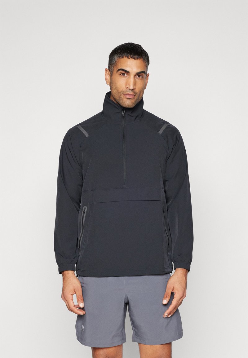 Zwarte pullover jack met een hoge kraag, halve rits, elastische manchetten en een borstzak. Gedeeltelijk gedragen met grijze shorts met subtiele accenten.