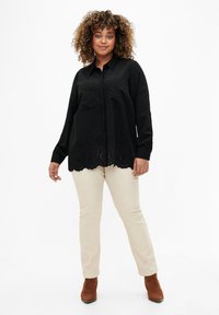 Camicia nera con bottoni, orlo scallop e ricami intricati nella parte inferiore, abbinata a pantaloni beige e stivaletti marroni alla caviglia.