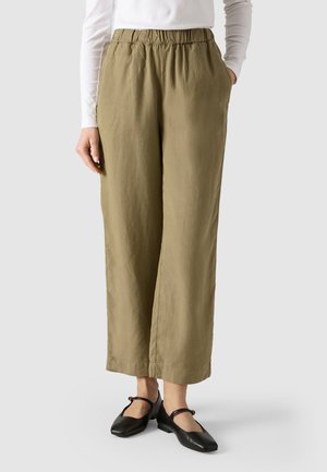 Woman wearing high-waist, olive green wide-leg pants with elastic waistband, white long-sleeve top, and black Mary Jane shoes on gray background.