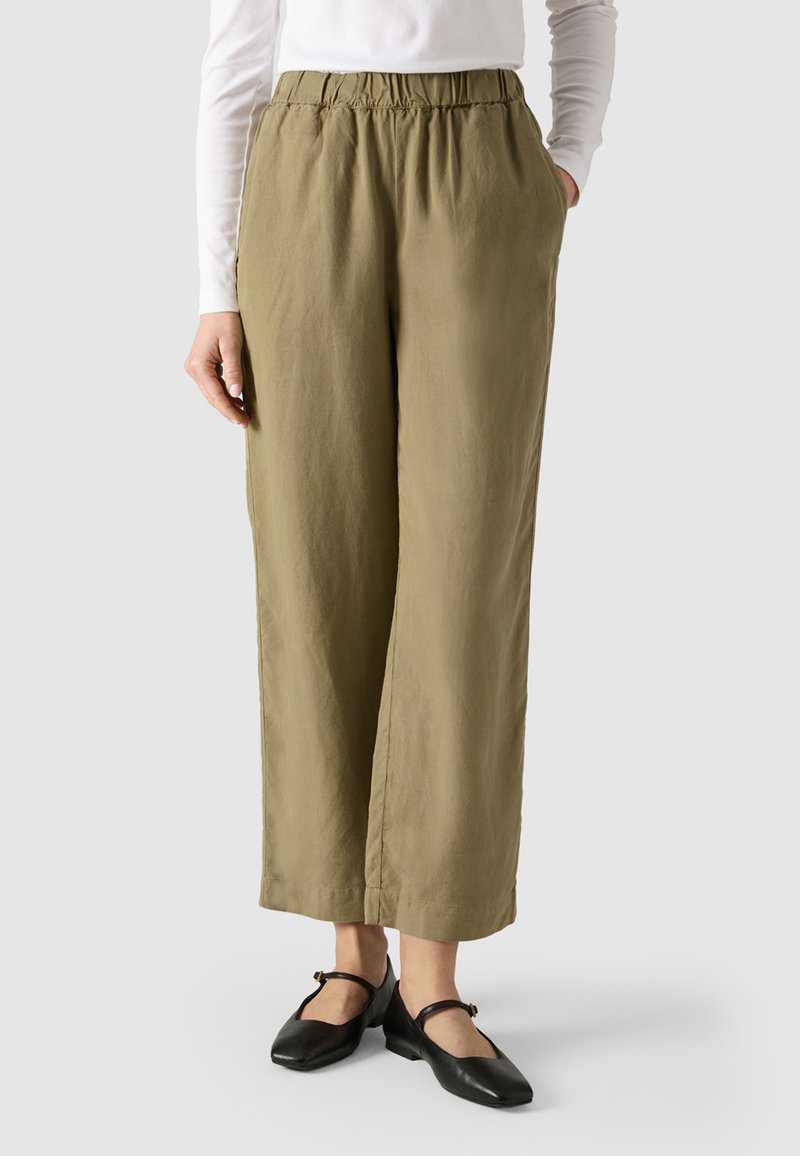 Kobieta ubrana w oliwkowo-zielone szerokie spodnie z wysokim stanem i elastycznym pasem, białą bluzkę z długim rękawem oraz czarne buty typu Mary Jane na szarym tle.