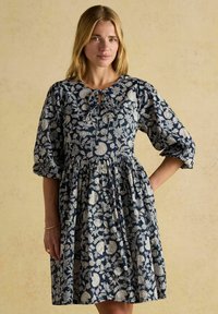 Eine Frau steht in einem navyblauen, blumigen Kleid mit Puffärmeln und einem Schlüsselloch-Ausschnitt und posiert vor einem sanften, neutralen Hintergrund.