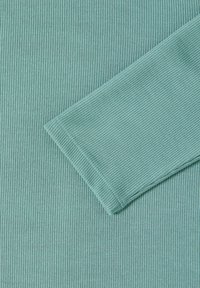 Minzgrüner Rippstoff mit glatter Textur. Mit langem Ärmelabschluss und sichtbaren Nähten entlang der Kanten für Langlebigkeit.