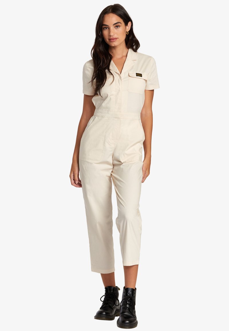 Cream-colored jumpsuit with short sleeves, front pockets, fitted waist, and tapered legs, paired with black ankle boots.