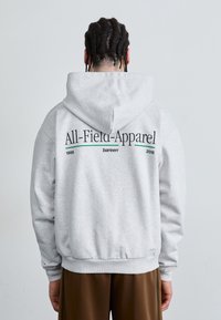 Grå hættetrøje med snor i hætten, med "All-Field-Apparel" trykt i sort og grøn på ryggen, ribbede manchetter og kant.