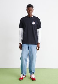 Black t-shirt with logo patch, layered over white long-sleeve shirt. Light blue jeans and red sneakers complete the look. Simple and casual design.