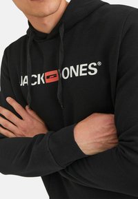 Sort hættebluse med snore, der har hvid tekst "JACK & JONES" og en rød detalje. Manchetten og kanten er med ribbet struktur. 100% bomuld.