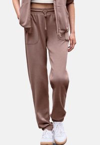Braune Jogginghose aus weichem Stoff, mit einem elastischen Bund und Kordelzug, seitlichen Taschen und eng anliegenden Bündchen. Tragen über weißen Sneakers.