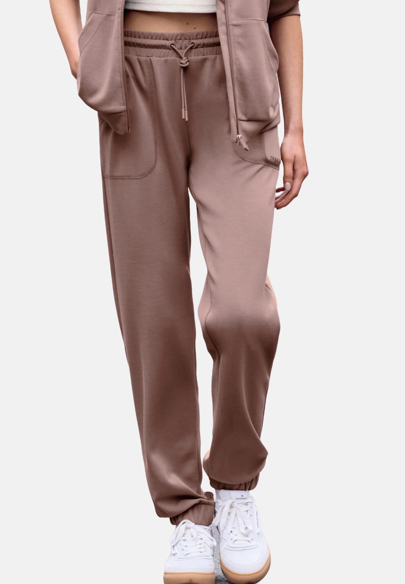 Braune Jogginghose aus weichem Stoff, mit einem elastischen Bund und Kordelzug, seitlichen Taschen und eng anliegenden Bündchen. Tragen über weißen Sneakers.