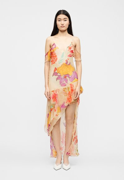 Femme portant une robe fluide et transparente à motifs floraux, avec des manches épaules dénudées et un ourlet asymétrique, associée à des chaussures blanches à bouts pointus.