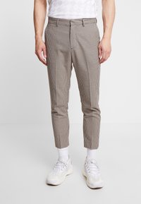 Hnědé kostkované kalhoty s úzkým střihem, s plochým předním dílem a olemovanými lemy. Kombinovány s bílými teniskami a kotníkovými ponožkami.