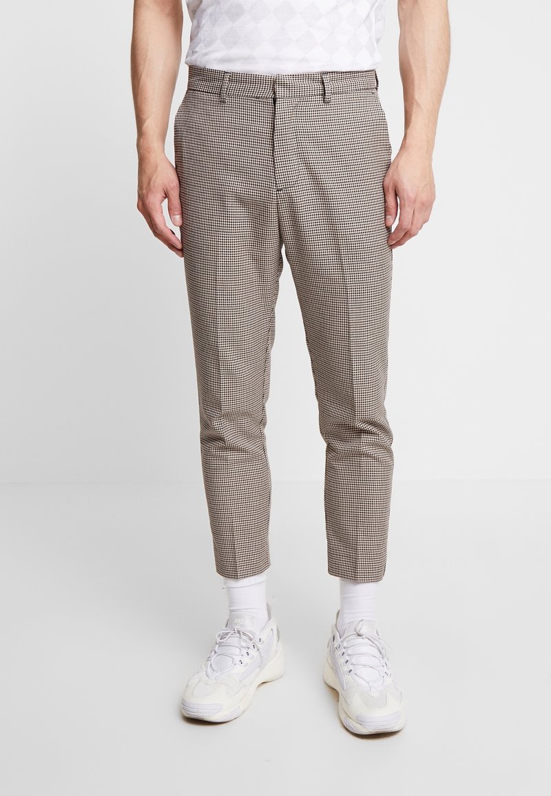 Hnědé kostkované kalhoty s úzkým střihem, s plochým předním dílem a olemovanými lemy. Kombinovány s bílými teniskami a kotníkovými ponožkami.