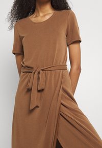 Robe courte marron à manches courtes en textile doux, avec un col rond et un détail de taille nouée. Présente une fente sur le côté pour un style supplémentaire.