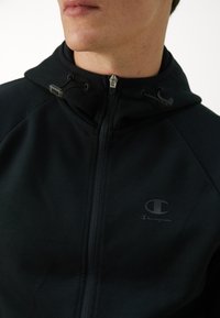 Schwarze Reißverschluss-Hoodie mit hohem Kragen, featuring ein Champion-Logo auf der Brust, verstellbaren Zugbändern und Raglanärmeln. Glatte Stofftextur.