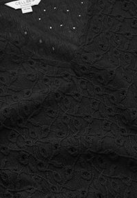 Tessuto nero con un intricato motivo in pizzo floreale e piccoli fori, che mostra dettagli di texture e cuciture, con un'etichetta di abbigliamento bianca.