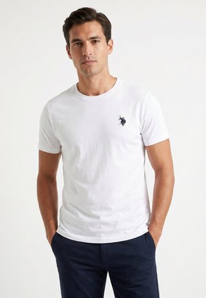 Homme portant un t-shirt blanc à manches courtes avec un petit logo sombre sur la poitrine et un pantalon foncé, debout les mains dans les poches.