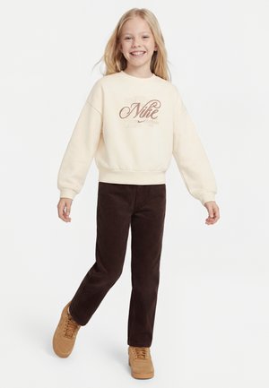 Copil zâmbitor care pozează, purtând un hanorac Nike crem, pantaloni din catifea maro închis și adidași bej, stând în picioare pe un fundal alb.