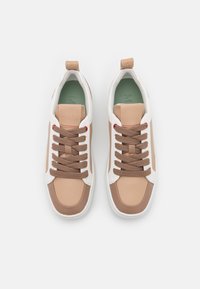 Baskets marron et beige avec une tige en cuir texturé, des accents blancs et des lacets marron. Comprend une semelle intérieure verte avec des détails de logo.