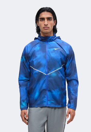 Homme aux cheveux foncés portant une veste zippée bleue Nike et un pantalon gris clair, debout face à l'avant sur un fond uni.