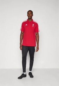 Červené polo tričko s bielymi pruhmi, s golierom a logom Liverpool FC. Čierne nohavice s bielymi bočnými pruhmi a čierne športové topánky.