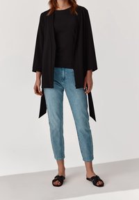 Svart öppen cardigan över en svart topp, kombinerad med ljusblå denimjeans och svarta knutna sandaler. Slät tygkvalitet med avslappnad passform.