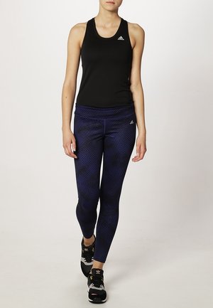 Mujer caminando, vistiendo una camiseta sin mangas negra de Adidas, leggings estampados azul oscuro y zapatillas negras de Adidas.