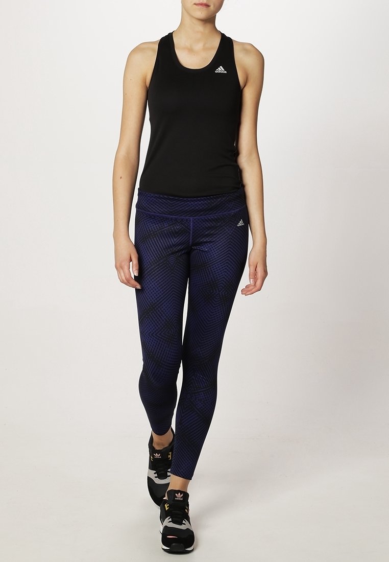 Femme marchant, portant un débardeur noir Adidas, des leggings bleu foncé à motifs, et des baskets noires Adidas.