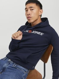 Un sweat à capuche bleu marine avec une poche avant, un texte logo blanc, un cordon de serrage pour la capuche et une texture douce, associé à un jean bleu clair sur une chaise en bois.
