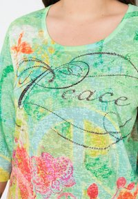 Grünes T-Shirt mit floralem Muster und weitem Rundhalsausschnitt, mit einem "Peace"-Schriftzug, verziert mit silbernen Nieten und aus strukturiertem Stoff.