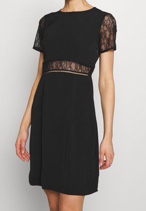 Kobieta nosząca krótką czarną sukienkę z krótkimi koronkowymi rękawami i koronkowym panelem wokół talii na neutralnym tle.