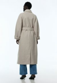 Beige wollen mixjas met lange mouwen, een ceintuur in de taille en opgerolde manchetten, gedragen over blauwe wijde jeans en zwarte high-heeled schoenen.