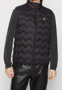 Czarna pikowana kamizelka z falistym wzorem, z wysokim kołnierzem i zamkiem błyskawicznym. Małe żółte logo znajduje się na lewym obszarze klatki piersiowej.