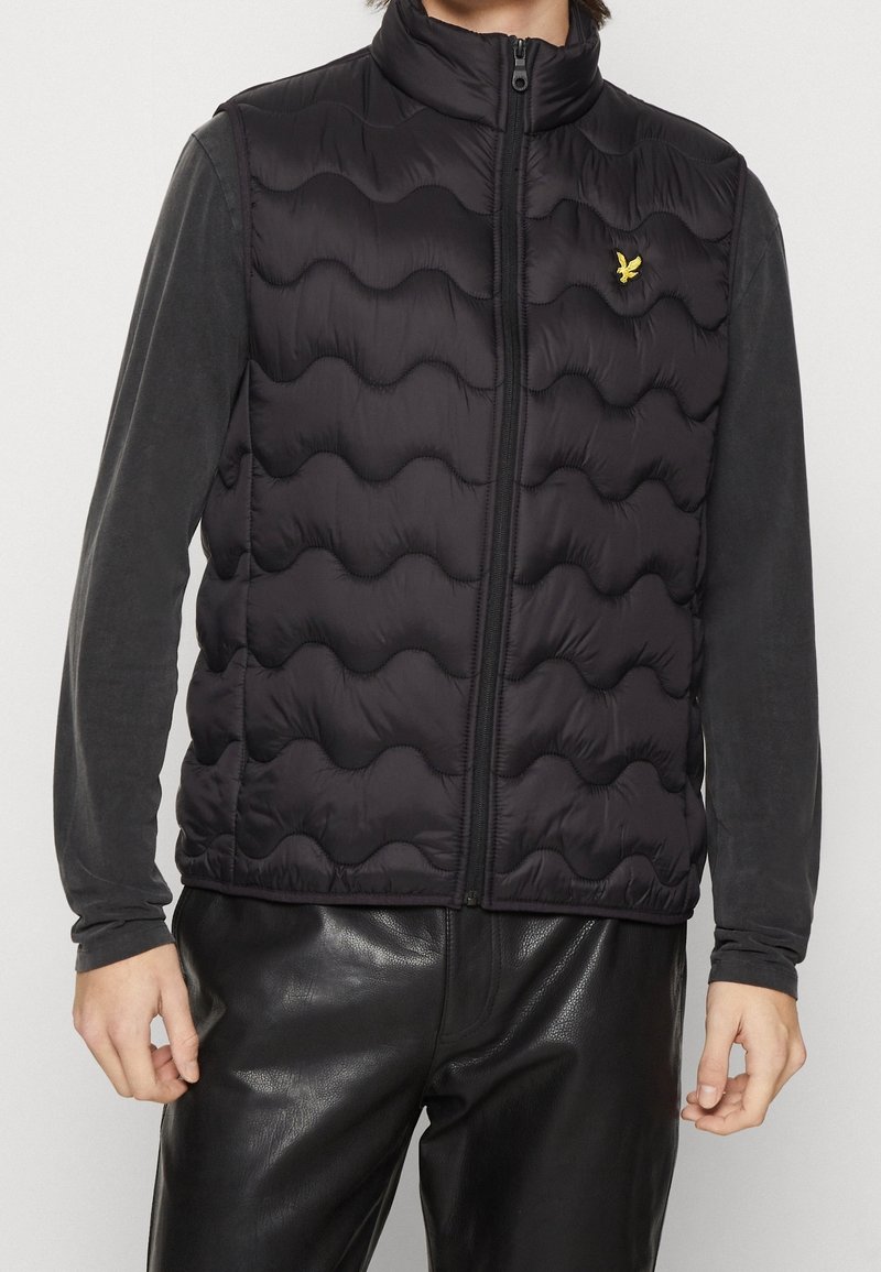 Czarna pikowana kamizelka z falistym wzorem, z wysokim kołnierzem i zamkiem błyskawicznym. Małe żółte logo znajduje się na lewym obszarze klatki piersiowej.