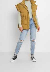 Chaleco acolchado sin mangas en color mostaza con un cuello alto, cierre de cremallera y bolsillos laterales, llevado sobre una camiseta verde menta y jeans desgastados.