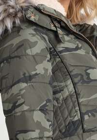 Olivengrøn pufferjakke med camouflage mønster, der har en faux fur-fleecelining i hætten, guld hardware og quiltede detaljer langs lynlåsen.