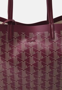 Borsa a tracolla bordeaux con superficie testurizzata. Caratterizzata da motivi geometrici e un logo Lacoste. Spallacci in pelle robusta per il trasporto.