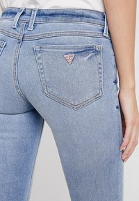 Jeans de mezclilla azul claro con un diseño ajustado, que presenta un pequeño logo triangular de Guess en el bolsillo trasero y sutiles detalles de desgaste.