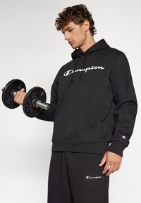 Hoodie en coton noir avec logo "Champion" blanc, poche avant et capuche avec cordon de serrage ; modèle tenant un haltère, complétant une tenue de sport.